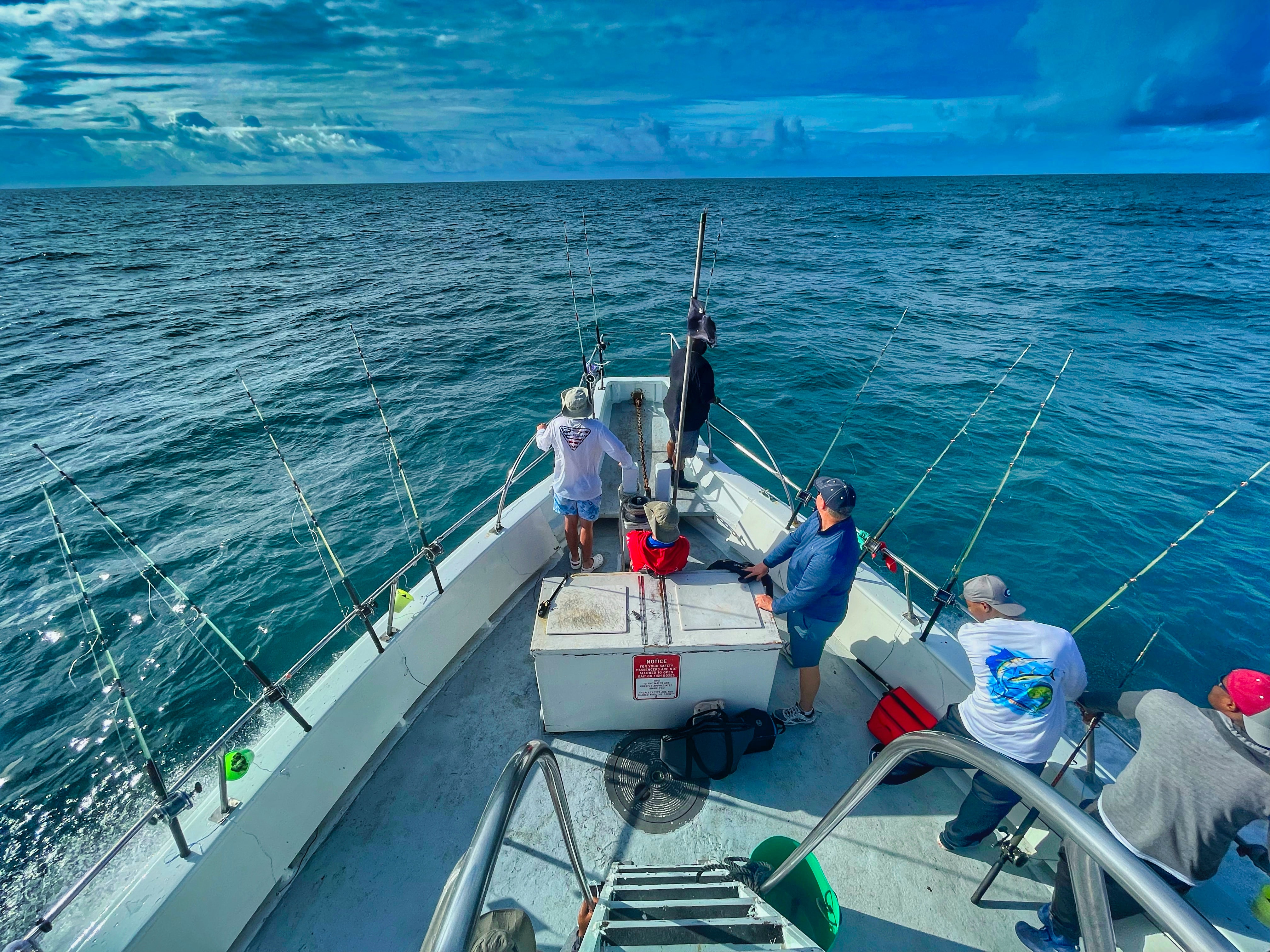 People fishing on a boat under a cloudy blue sky with rods lined up.